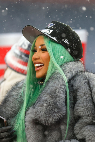 Cardi B wears a gray fur coat and mint green wig at a football game.